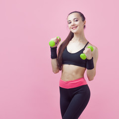 A young athletic woman, with a smile, in black sports top, doing aerobics on pink background with green light dumbbells.