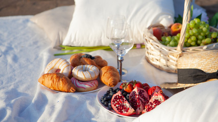 Picnic in the rays of the setting sun on the sand. On the plaid lies basket of fruit, glasses and sweet pastries.
