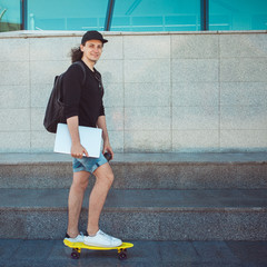 A young man with a laptop under his arm stands on the background of a gray wall, putting one foot...
