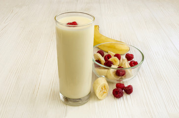 Banana yogurt in a tall glass and raspberries in a cup on a white wooden background