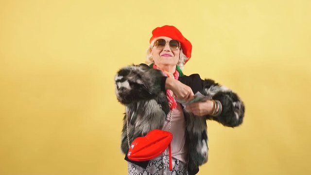 Rich elegant woman in fur coat and red cap throwing money, spending money on useless thing, isolated yellow background. studio shot