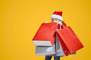 Adorable child in red Santa hat and sweater with deer carrying colorful shopping bags with Christmas gifts on yellow background