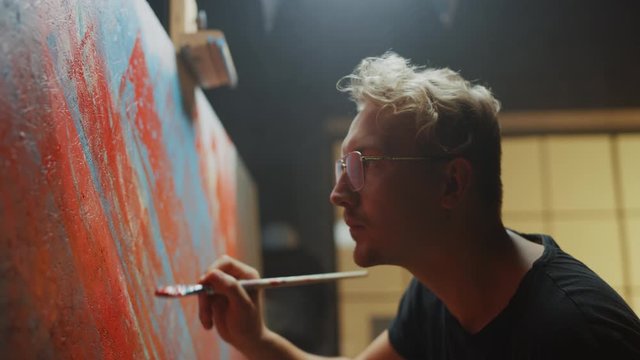 Portrait of Artist Working on Abstract Painting, Uses Paint Brush To Create Daringly Emotional Modern Picture. Dark Creative Studio Large Canvas Stands on Easel Illuminated. Side View Close-up Shot 