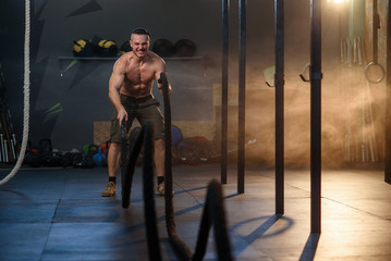 Handsome strong man doing exercises for the muscles with battle ropes in fitness gym.