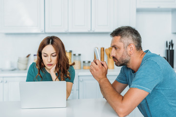 Obraz premium husband and wife sitting at kitchen table and using smartphones