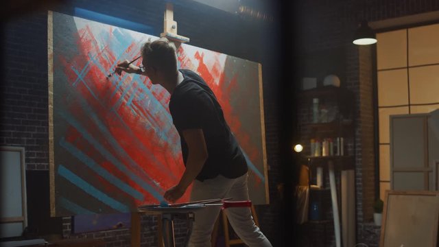 Talented male Artist Works on Abstract Oil Painting, with Broad Strokes of Paint Brush he Creates Modern Masterpiece. Dark and Messy Creative Studio where Large Canvas Stands on Easel Illuminated 