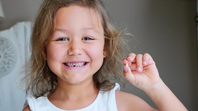 Portrait of cute little cheerful child girl is showing her lost milk tooth and smiling to camera of toothless mouth while she standing in bedroom at home. Happy baby holding her fallen tooth in hand