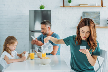 woman with smartphone overfilling bowl with milk while sitting at kitchen table near husband and daughter using smartphones