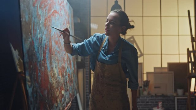 Female Artist Works on Abstract Oil Painting, Moving Paint Brush Energetically She Creates Modern Masterpiece. Dark Creative Studio where Large Canvas Stands on Easel Illuminated. Zoom Out Shot