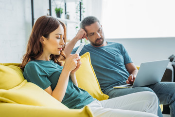 thoughtful man with laptop sitting on sofa near wife using smartphone at home