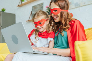 happy woman with daughter in costumes of superheroes using laptop at home