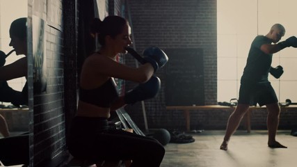 Beautiful Fit Brunette Kickboxer Athlete Sitting on a Bench in Loft Gym with Motivational Posters. She's Putting on Her Blue Fighting Gloves. Woman Stands Up and Continues Her Intense Boxing Training.