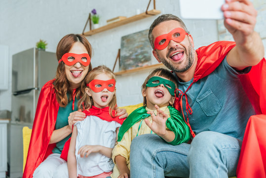 selective focus of happy family in superheroes costumes sticking tongues out while taking selfie on smartphone at home
