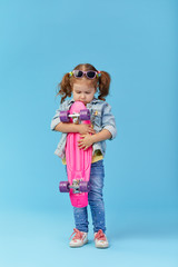 Cool little toddler girl in denim clothes holds a skateboard and funny looking at the camera isolated on blue background