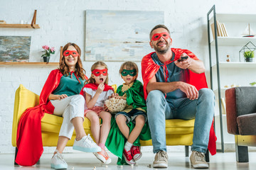 happy family in costumes of superheroes watching tv while sitting on sofa with popcorn