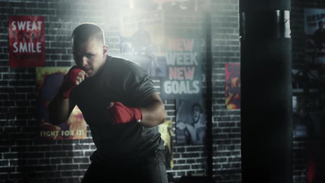 Strong and Handsome Male Boxer is Working Out in a Loft Gym with Motivational Posters on Walls. Masculine Athlete is Practising Punches in the Air. He's Serious and Energetic. Room is Dark.