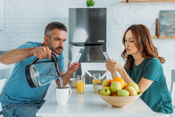 man with smartphone pouring water in cup while sitting in kitchen near wife using smartphone