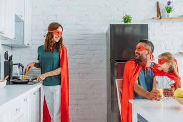 family in coustumes of superheroes spending time in kitchen, while father and daugher drinking orange juice and mother preparing breakfast