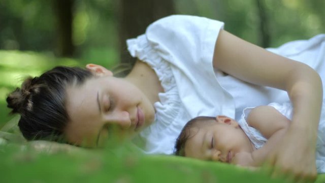 Mother kisses her Baby sleeping on a green grass Outdoors. Happy young Mother and Child in Green Summer Park. Beautiful family in spring park enjoying nature.