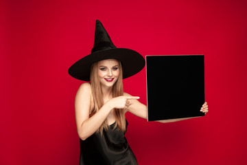 Young blonde woman in black hat and costume on red background. Attractive caucasian female model posing. Halloween, black friday, cyber monday, sales, autumn concept. Copyspace. Holds black plate.