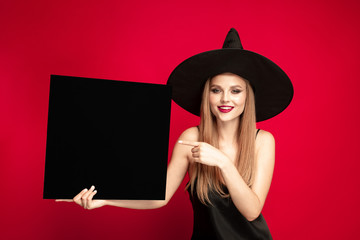 Young blonde woman in black hat and costume on red background. Attractive caucasian female model posing. Halloween, black friday, cyber monday, sales, autumn concept. Copyspace. Holds black plate.