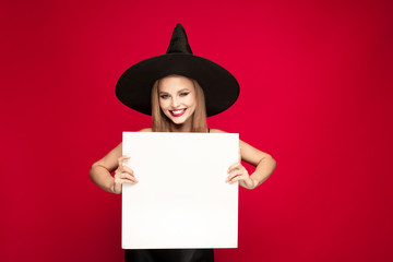 Young blonde woman in black hat and costume on red background. Attractive caucasian female model posing. Halloween, black friday, cyber monday, sales, autumn concept. Copyspace. Holds white plate.