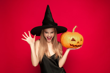 Young blonde woman in black hat and costume on red background. Attractive caucasian female model. Halloween, black friday, cyber monday, sales, autumn concept. Copyspace. Holds pumpkin, screams.