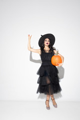 Young brunette woman in black hat and costume on white background. Attractive caucasian female model. Halloween, black friday, cyber monday, sales, autumn concept. Copyspace. Holds pumpking, pointing.