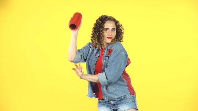 Girl listening to music with bluetooth portable speaker and dancing on yellow background at studio.