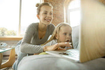 Female caucasian teacher and little girl, or mom and daughter. Homeschooling. Sitting on the sofa and using laptop for knowledges getting while lesson is. Education, school, studying concept.