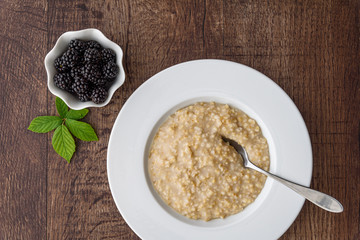 Cooked oatmeal in a white bowl with a metal spoon, small side bowl of fresh blackberries, on a wood table