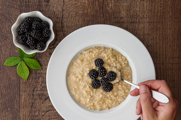 Cooked oatmeal in a white bowl with hand holding a metal spoon, small side bowl of fresh blackberries, on a wood table