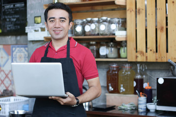 close up asian man barista smiling holding laptop checking orders and stock behind the counter. cafe owner standing in restaurant