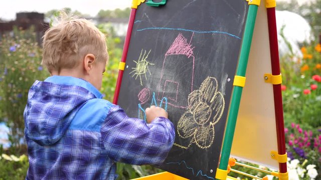 Young artist draws on the Board. A boy in the open air among the flowers and greenery draws children's pictures with chalk on the Board.