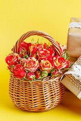 Wicker basket with roses and beautifully wrapped gifts on yellow background.