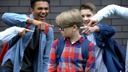 Schoolchildren pointing fingers at junior student, mocking smart boy, bullying