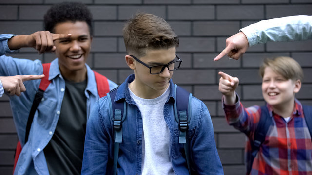 Cruel schoolchildren pointing fingers at student, mocking smart boy, bullying