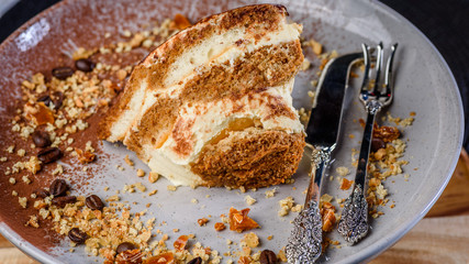 A slice of Tiramisu on a plate surrounded by coffee beans, honeycomb and nuts and some fancy cutlery