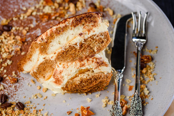 A slice of Tiramisu on a plate surrounded by coffee beans, honeycomb and nuts and some fancy cutlery