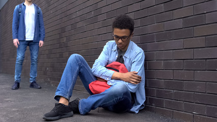 Bullied afro-american schoolboy sitting on ground, scared of offender behind