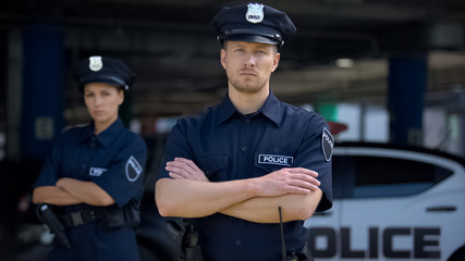 Confident male and female police officers in uniform standing near patrol car