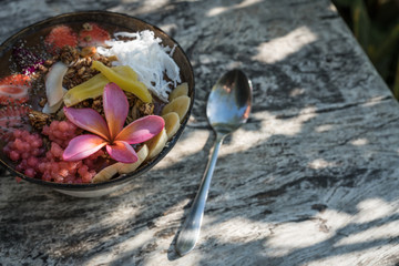 smoothie bowl in a coconut shell plate with with strawberries, chia seeds, banana, granola, flower on wooden background. Green tropical leaves background