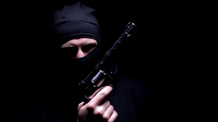 Dangerous man in balaclava mask holding gun, black background, serial killer