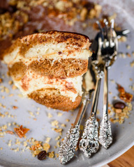 A slice of Tiramisu on a plate surrounded by coffee beans, honeycomb and nuts and some fancy cutlery