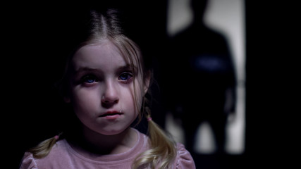 Little crying girl looking at camera, silhouette of rude father outside door