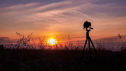 Silhouette of camera on tripod capturing sunset over the wheat field. Photographing an evening landscape. Camera the night view.