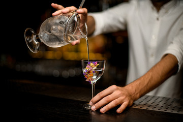 Male bartender pouring a cocktail from the measuring cup with a strainer to a flower decorated glass