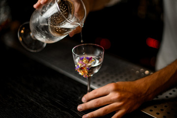 Male bartender pouring a cocktail from the measuring cup with a strainer to a glass decorated with flowers