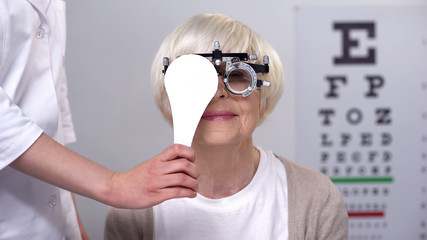 Ophthalmologist closing eye to senior lady in phoropter, checking lens diopter