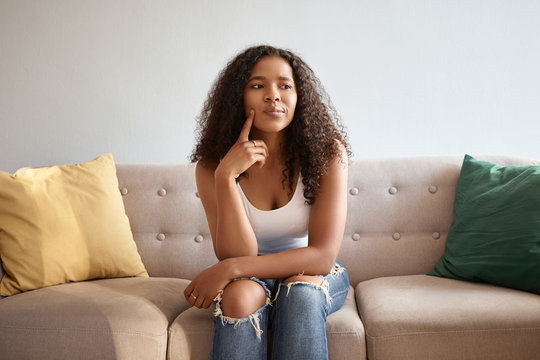 Indoor image of fashionable cute girl with African appearance sitting isolated on gray sofa with yellow and green cushions having doubtful thoughtful look, touching chin, saying Hmm, Let me think